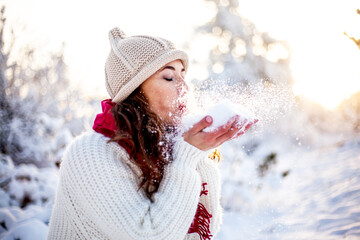 Happy woman walking in the snowy winter day outdoor. Female mdel dressed white sweater and hat blowing snow and posing against the  sun