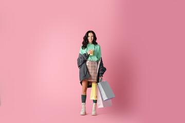  brunette young woman in casual winter outfit with shopping bags and coffee to go on pink background