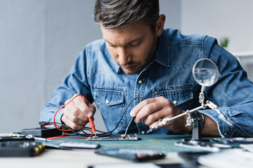 Focused repairman holding sensors of multimeter on disassembled part of mobile phone on blurred foreground