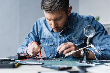 Focused repairman holding sensors of multimeter on disassembled part of mobile phone on blurred foreground