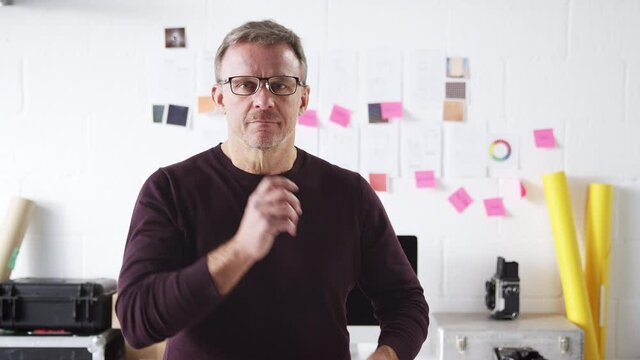 Portrait of mature businessman sitting in front of desk in studio of start up fashion business putting on glasses and smiling at camera- shot in slow motion
