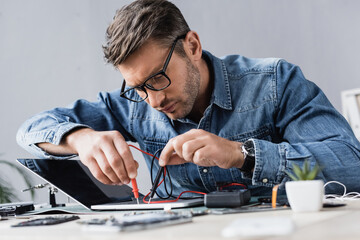 Focused repairman holding sensors of multimeter on broken keyboard of laptop at workplace on...