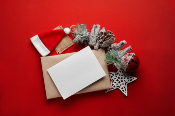 Christmas composition. The concept of winter, new year. Flat lay, top view, space for text. Blank card on gift box made of kraft paper with hat, spruce branches and toys on red table with snow