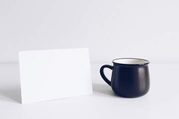 Enamel black mug and horizontal postcard on white table background mockup. Boho style classic stock photo. Still life composition with black ceramic cup. Hygge scene, product mock up template