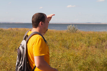 Fototapeta premium A man in an orange t-shirt with a backpack looks out to sea. The traveller shaded his eyes with his hand.