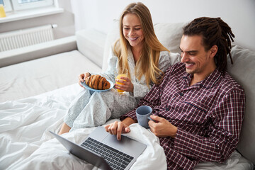 Happy young couple using laptop and having breakfast in bed