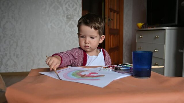 Caucasian boy artist in children room sits at the table with art supplies paints, takes brush and draws.