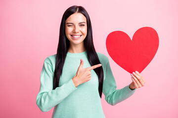 Photo portrait of winking girl pointing finger at big red heart card in one hand isolated on pastel pink colored background