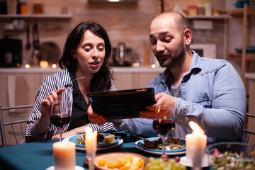 Cheerful young couple dining together using tablet during romantic dinner. Adults sitting at the table in the kitchen browsing, searching, , internet, celebrating, anniversary, happy., festive, enjoy.