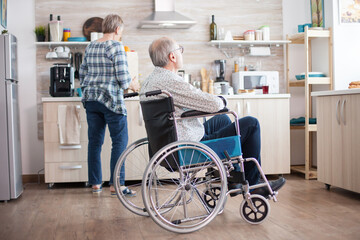 Pensive disabled man in wheelchair looking through kitchen window while wife is unpacking food from...