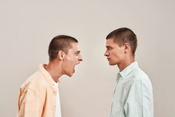 Portrait of two young caucasian men, twin brothers arguing, shouting while standing face to face isolated over beige background