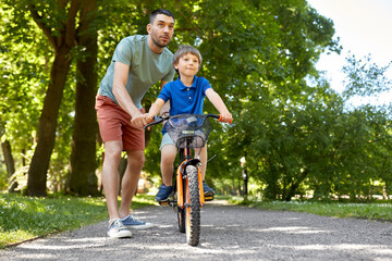 family, fatherhood and leisure concept - happy father teaching little son to ride bicycle at park