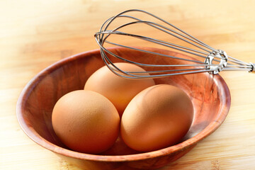 Three fresh brown eggs in a bamboo bowl and a wire whisk on a wooden table. Concept of...