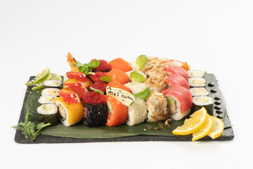 Large assortment of assorted sushi maki made from salmon, tuna, shrimp roe of tobiko flying fish, rice and sesame seeds on a black slate stone plateau, garnished with bamboo leaves, teriyaki sauce.