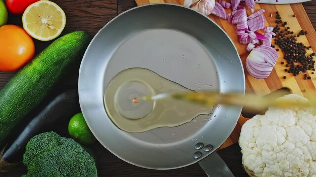 Cook pours sunflower oil into a frying pan, top view. Kitchen pan stands on brown boards, vegetables around it, and oil is poured into it for frying.  Slow motion  of  oil pouring into a frying pan. 
