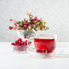 Autumn red herbal tea in transparent cup, spoon with hawthorn, rosehip berry, dried leaves in sun beam in soft light white interior, square.