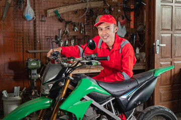 Fototapeta na wymiar pose of young mechanic in wearpack uniform and hat standing beside a dirt bike in garage