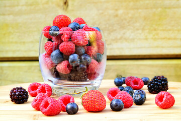 A glass full of fresh strawberries, raspberries, blueberries, and blackberries with more berries around it on a wooden table.