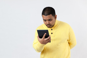 Image of amazing serious asian man wearing yellow shirt using tablet computer. Looking aside.
