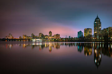 Obraz premium Night view of modern buildings in the Obolon district of Kiev, Ukraine, close to the Dnieper River, The lights reflect on the calm water.