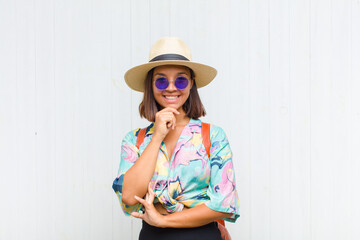 latin woman looking happy and smiling with hand on chin, wondering or asking a question, comparing...
