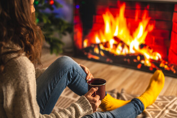 Girl in a sweater and socks is sitting next to the fireplace with cup of beverage. Concept of creating a cozy atmosphere
