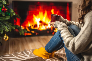 Girl in a sweater and socks is sitting next to the fireplace with cup of beverage. Concept of creating a cozy atmosphere