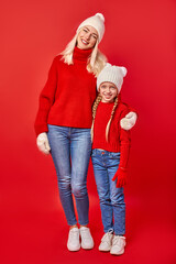 mother woman and child girl in warm clothes posing at camera, full-length portrait, isolated on red studio background