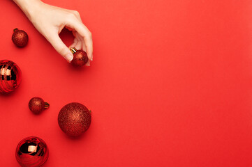 Red Christmas balls on a red background top view. A female hand holds a festive New Year's decor, flat lay. Xmas winter decorations to celebrate the seasonal holiday. Balls with glitter and sparkles.