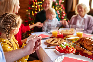 family members praying before eating, sitting together behind served dinner table, celebrating thanksgiving day and easter. focus on hands