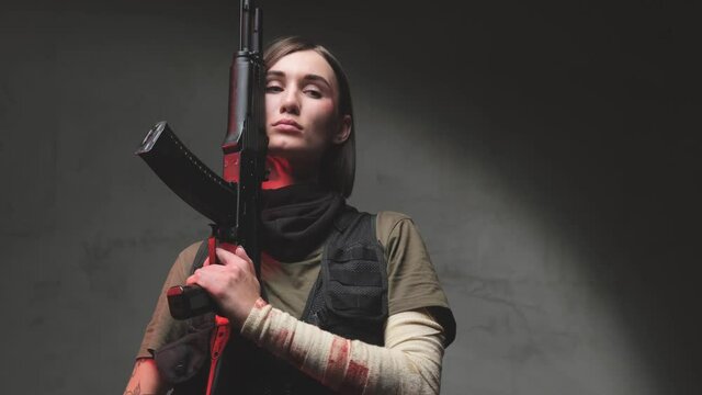 Soft and seductive martial woman with short haircut and dressed in black vest with dark shirt poses in dark background holding a rifle near her face with her bandaged arm.