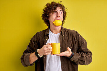 young curly caucasian male eating lemon for being healthy, eat it with tea, hold cup in hands,...