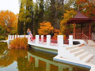 Redhead girl sits on a bridge over a green muddy pond in a park against the background of autumn trees and a gazebo