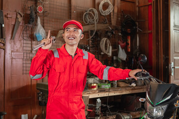 pose of a mechanic finding a good idea wearing a wearpack and a hat carrying a wrench near a dirt bike at the garage