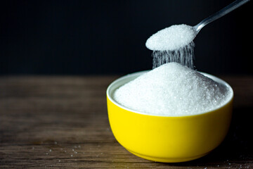 A picture of white sugar piled up in a yellow bowl with a spoon pouring sugar down on it.A black background as a very sweet food or seasoning may be at risk of diabetes.