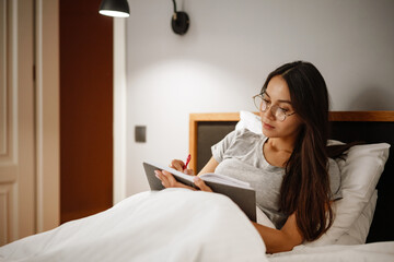 Serious beautiful woman writing down notes while sitting in bed