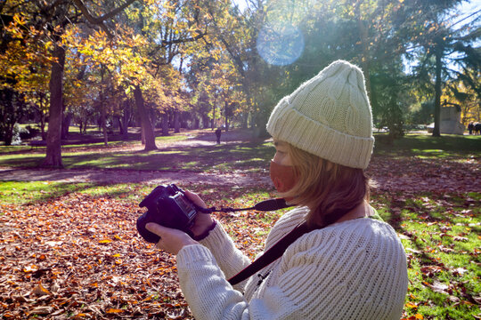 Woman wearing a mask, sweater and white hat, checking her photos on camera, in an autumnal park, in the morning, horizontal