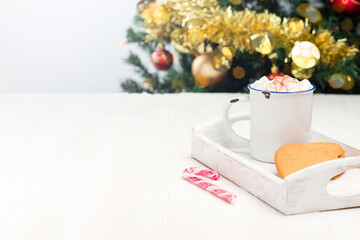 Heart shaped gingerbread cookies or pepparkakor with cup of hot chocolate and candy cane, on white wooden tray with christmas tree on background, horizontal, copy space