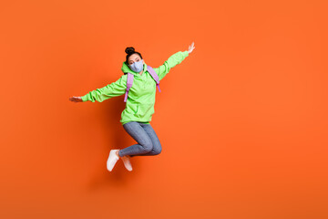 Fototapeta na wymiar Full length photo portrait of schoolgirl jumping up making plane with hands wearing blue fabric face mask isolated on vivid orange colored background
