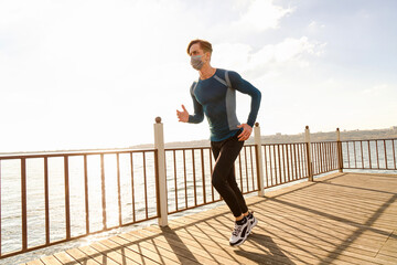 healthy and handsome young male model running on the beach. The man wearing a mask is running by the sea in sportswear. Healthy man who does sports outside with his mask due to corona virus measures.