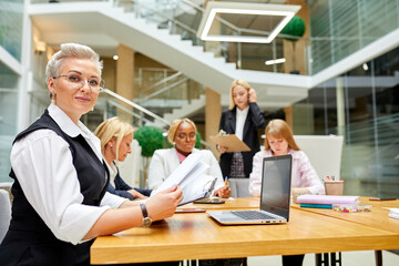 Obraz premium attractive business lady in the office, multi-ethnic group of co-workers in the background. she looks at camera smiling