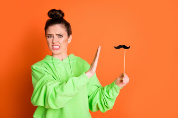 Photo of confused young woman wear green hoodie not want use stick mustache isolated vivid orange color background