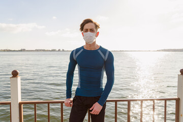 Handsome caucasian male in sportswear poses on the beach wearing a mask. young sportsman male walks outside at sunset by the sea in his protective mask.