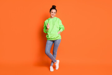 Fototapeta na wymiar Full size photo portrait of chill girl pulling hoodie string with hand in pocket crossed legs isolated on vivid orange colored background