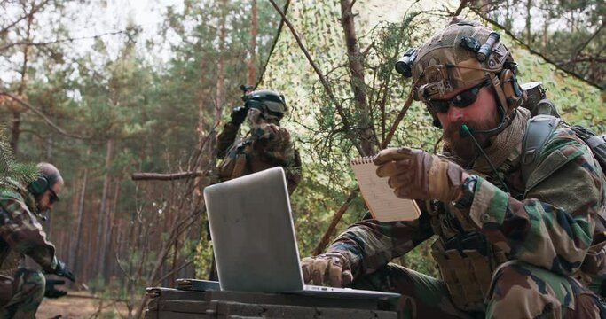A bearded brigade commander in a military uniform, with a helmet on his head, checks the coordinates from his official notebook and analyzes them on a military laptop.