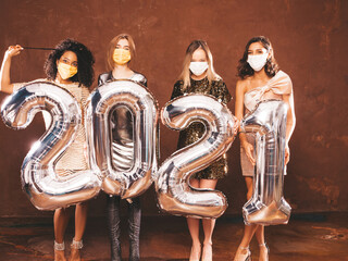 Beautiful Women Celebrating New Year.Happy Gorgeous Female In Stylish Sexy Party Dresses Holding Silver 2021 Balloons, Having Fun At New Year's Eve Party In Studio. In Medical Protective Masks