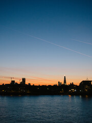 view of skyline over london buildings in silhoette the sky is a deep blue with orange red on the horizon with the river in the foreground, there are plane cloud streaks in the air with a tinge to them