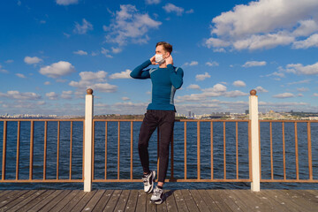 Male model posing with mask by the sea on a sunny autumn day. Image of handsome caucasian young man wearing a mask under blue sky with sportswear. Avaible copy space area. 