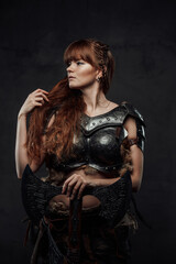 Fototapeta na wymiar Beautiful but dangerous warlike woman viking with long brown hairs in dark armour holding huge axe in dark background.