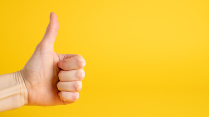 Hand thumbs up sign on yellow background. Approval gesture. Left side. Closeup of hand showing thumbs up sign